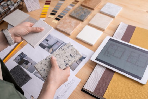 Hands of young female designer holding two samples of marble tile over wooden table with digital tablet, photos of home interior etc